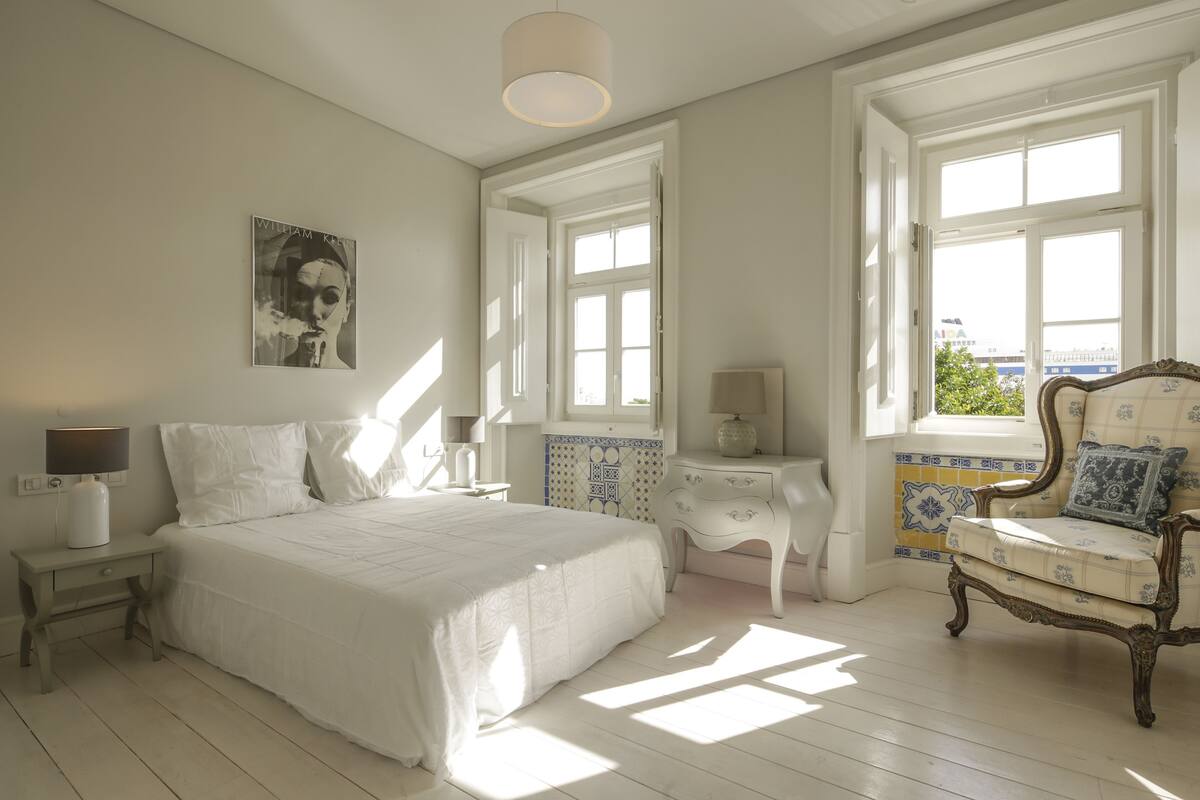 A bedroom with a large bed, a chair, a nightstand, a lamp, and a window with a view.