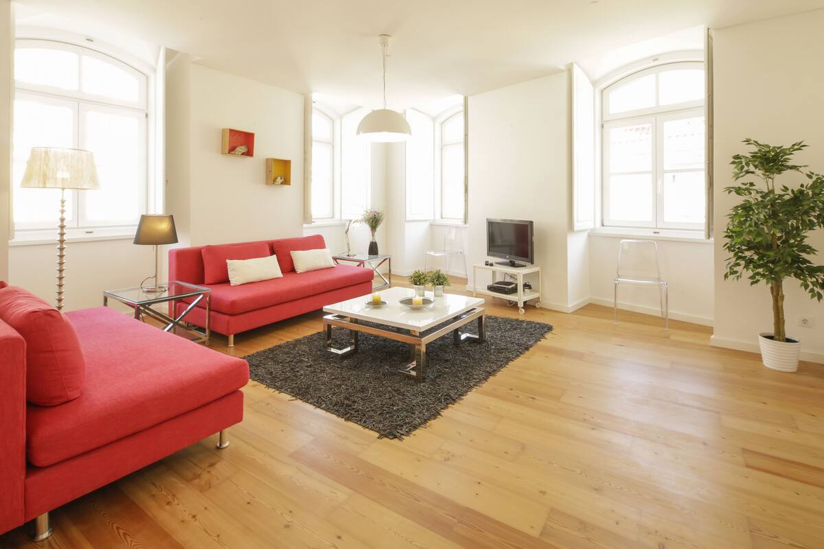 A spacious living room with a red sofa, a coffee table, and a TV. There are large windows providing natural light and a potted plant.