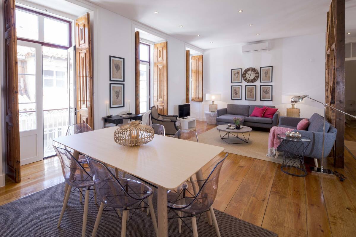 A spacious living room with a large wooden table, clear chairs, a grey sofa, and a coffee table.