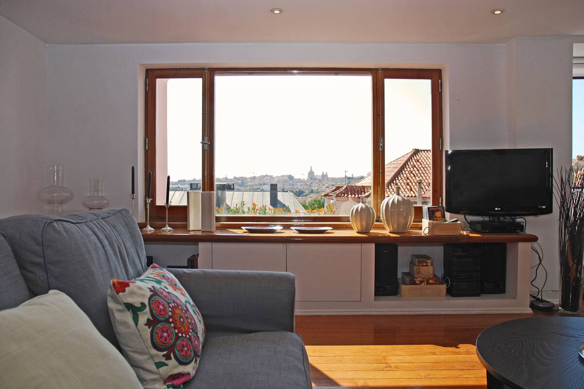 A living room with a large window, a grey sofa, a wooden coffee table, and a television on a stand.