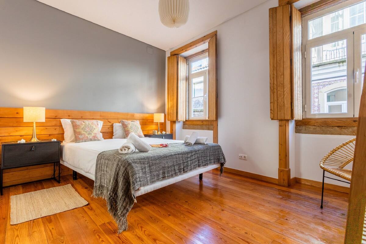 A bedroom with a wooden bed, nightstands, a chair, and large windows.