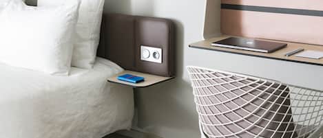 Una habitación de hotel moderna con una cama, un escritorio con una computadora portátil, una silla y una mesita de noche con una lámpara.