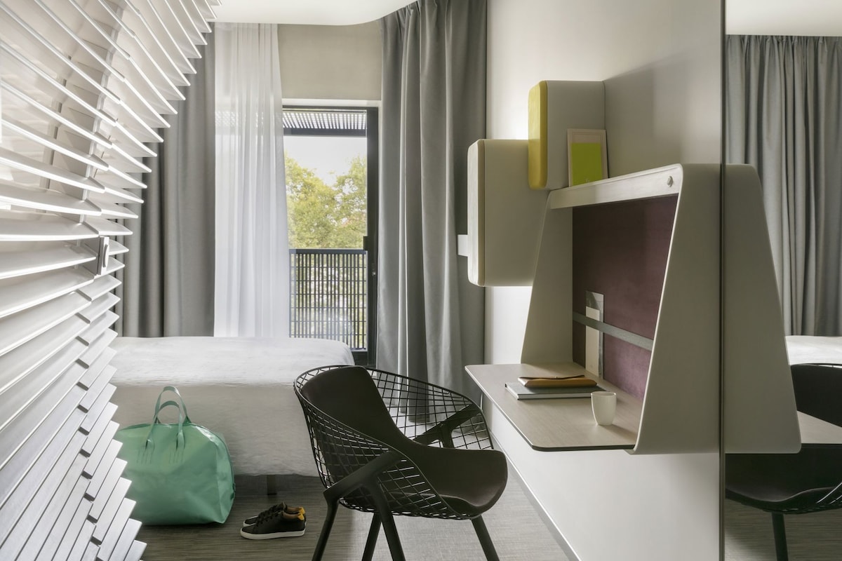 Una habitación de hotel moderna con cama, escritorio, silla, bolso y ventana con persianas.
