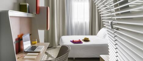 A modern hotel room with a bed, a desk with a laptop, a chair, and a window with blinds.