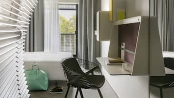 Una habitación de hotel moderna con una cama, un escritorio con computadora, una silla, un ventanal con cortinas y un bolso en el suelo.