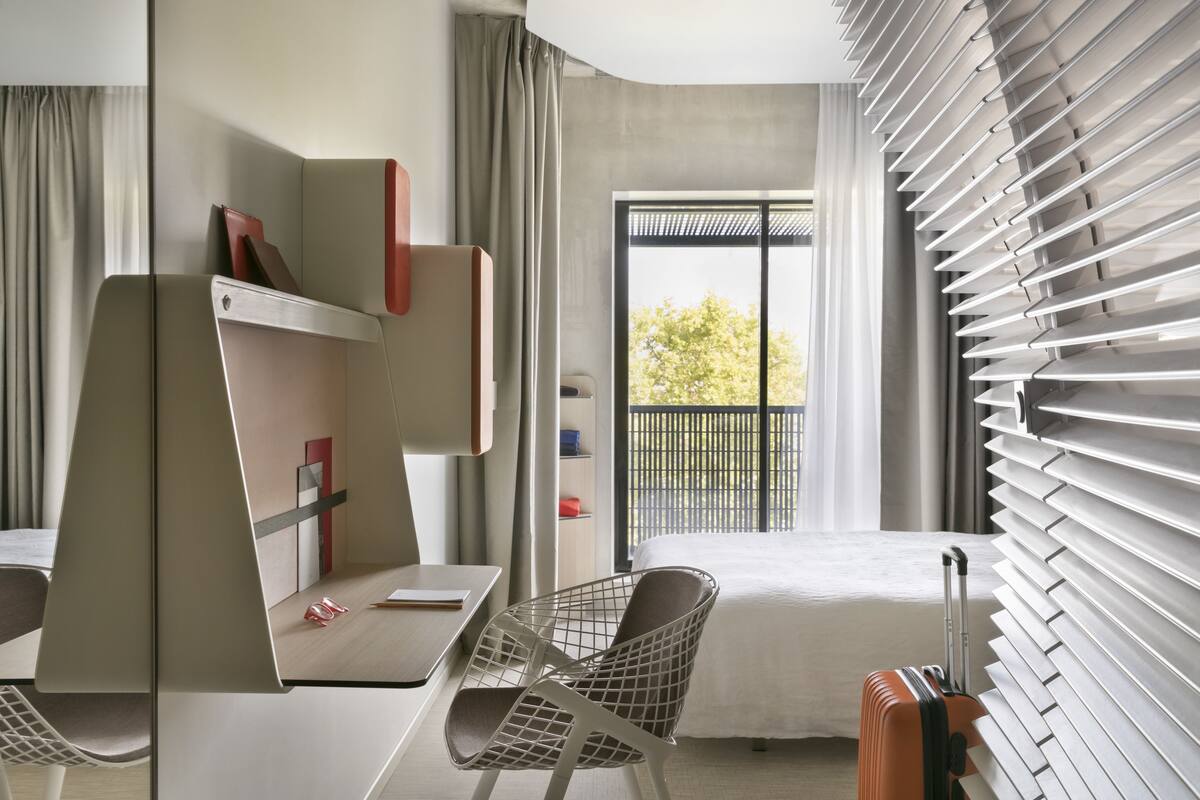 A modern hotel room with a bed, a desk, a chair, and a suitcase.
