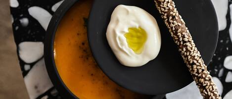 A bowl of soup with a dollop of cream and a sesame seed-covered breadstick on a black plate, set on a terrazzo floor.