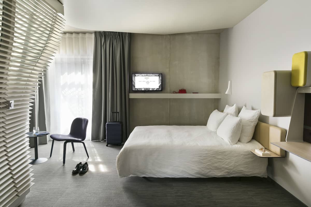 A hotel room with a bed, a TV mounted on the wall, a chair, a desk, and a suitcase.