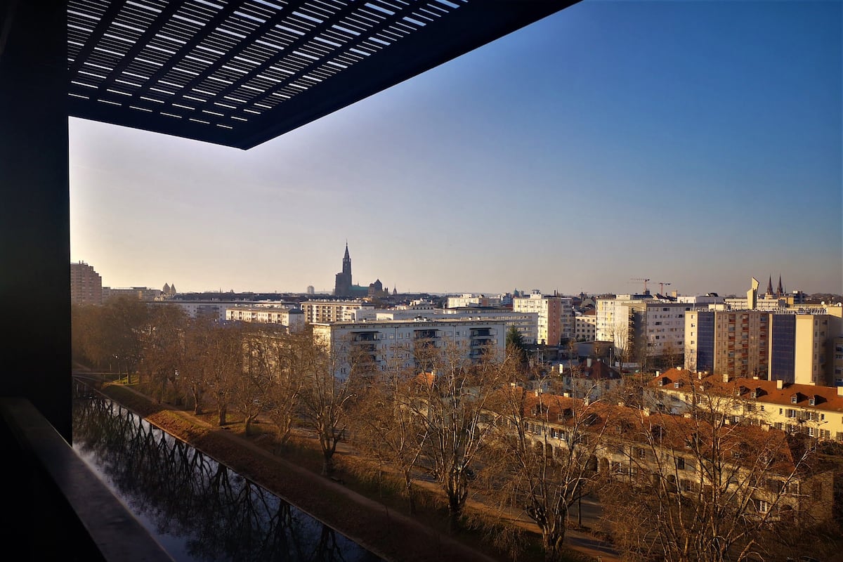 A cityscape with a prominent church spire, modern buildings, and a bridge with trees in the foreground.