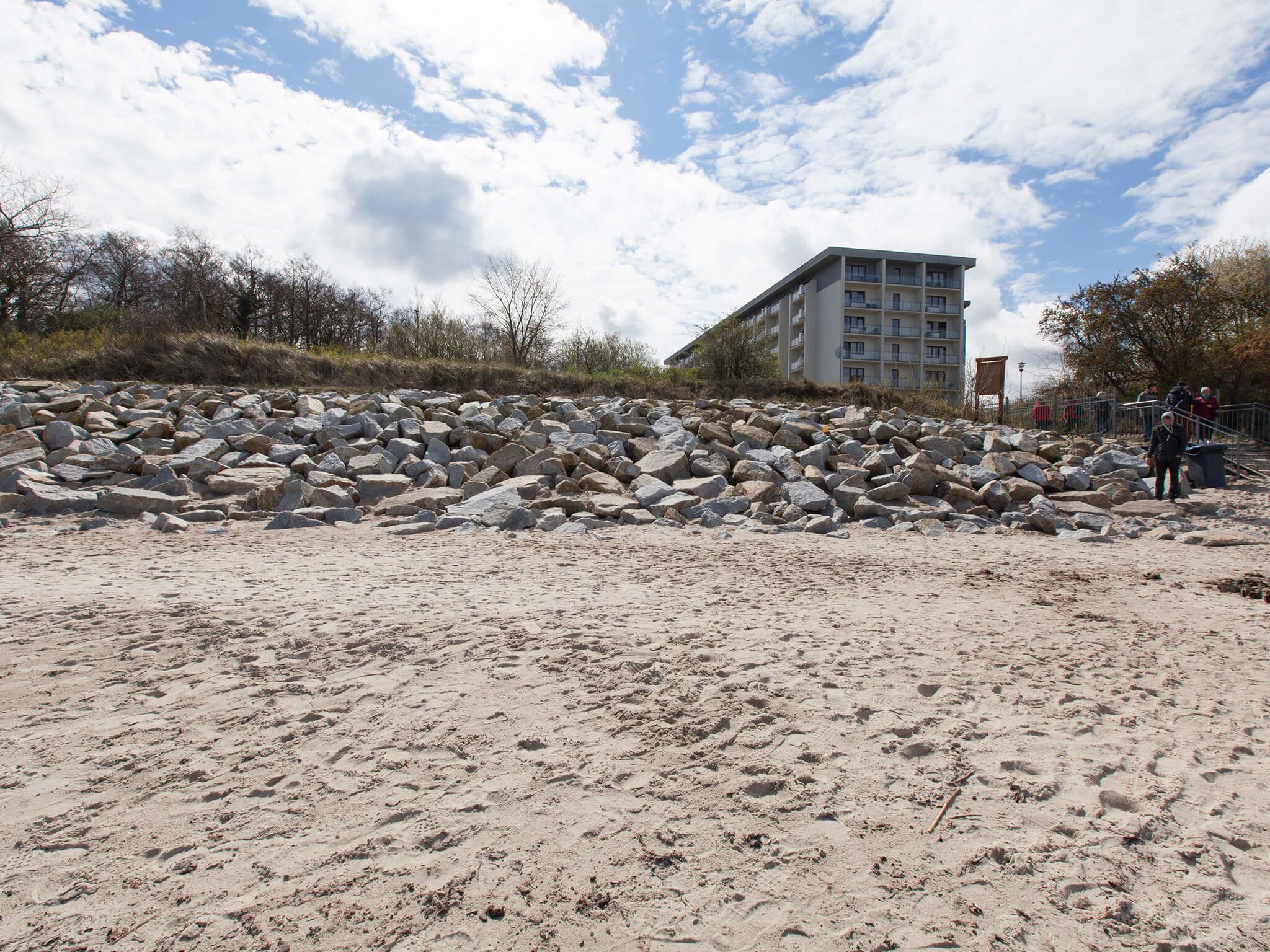 A sandy beach with large rocks in the foreground and a building with multiple balconies in the background.