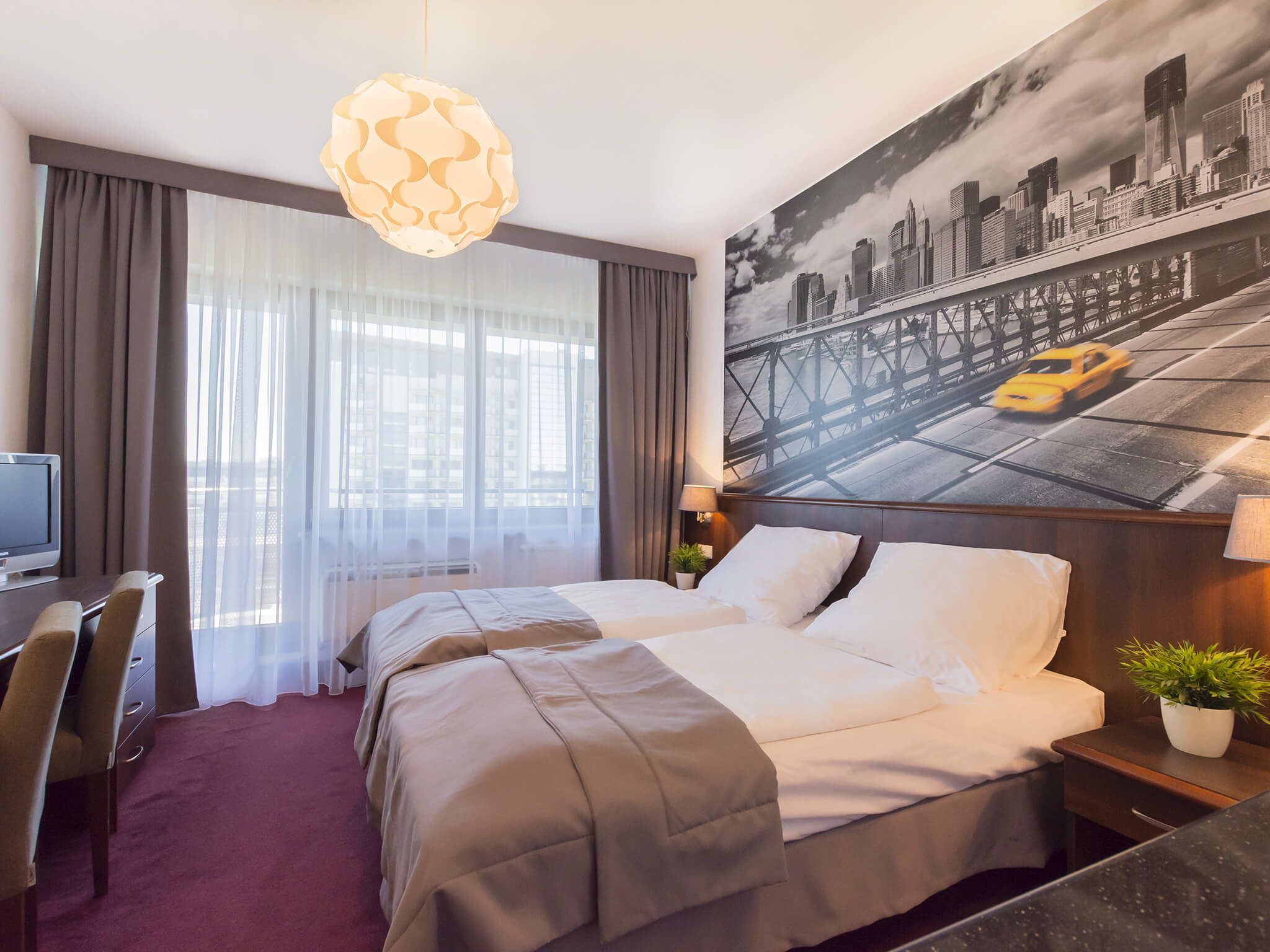 Ett hotellrum med en säng, ett skrivbord, en stol, en tv, ett stort fönster med tunna gardiner, en hängande lampa och en väggmålning som föreställer en stadsbild.