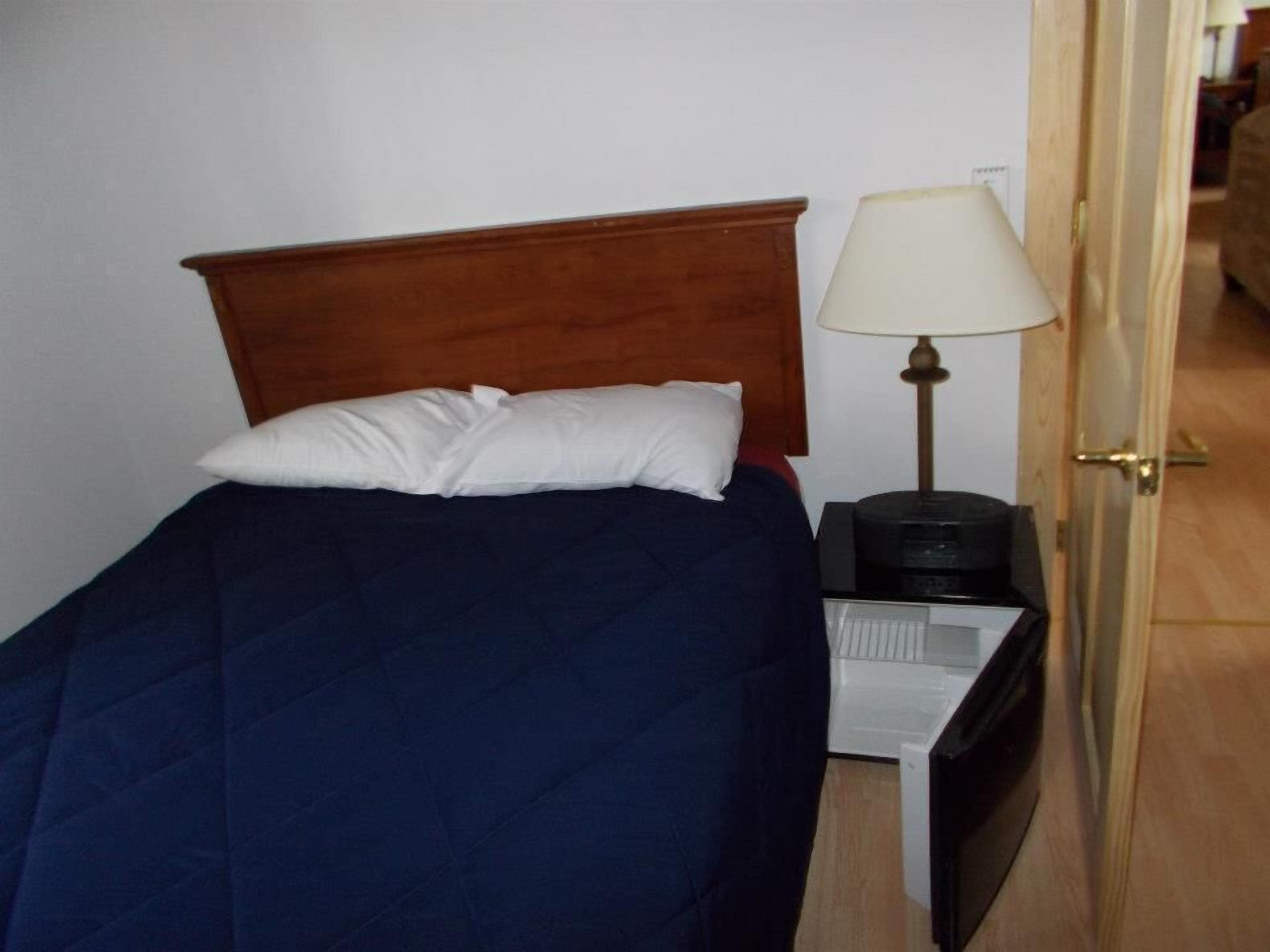Un letto singolo con una testiera in legno, un cuscino bianco e una coperta azzurra. Un comodino con una lampada e un oggetto nero, forse un telefono o un telecomando, e una scatola contenitore bianca visibile sotto il tavolo.