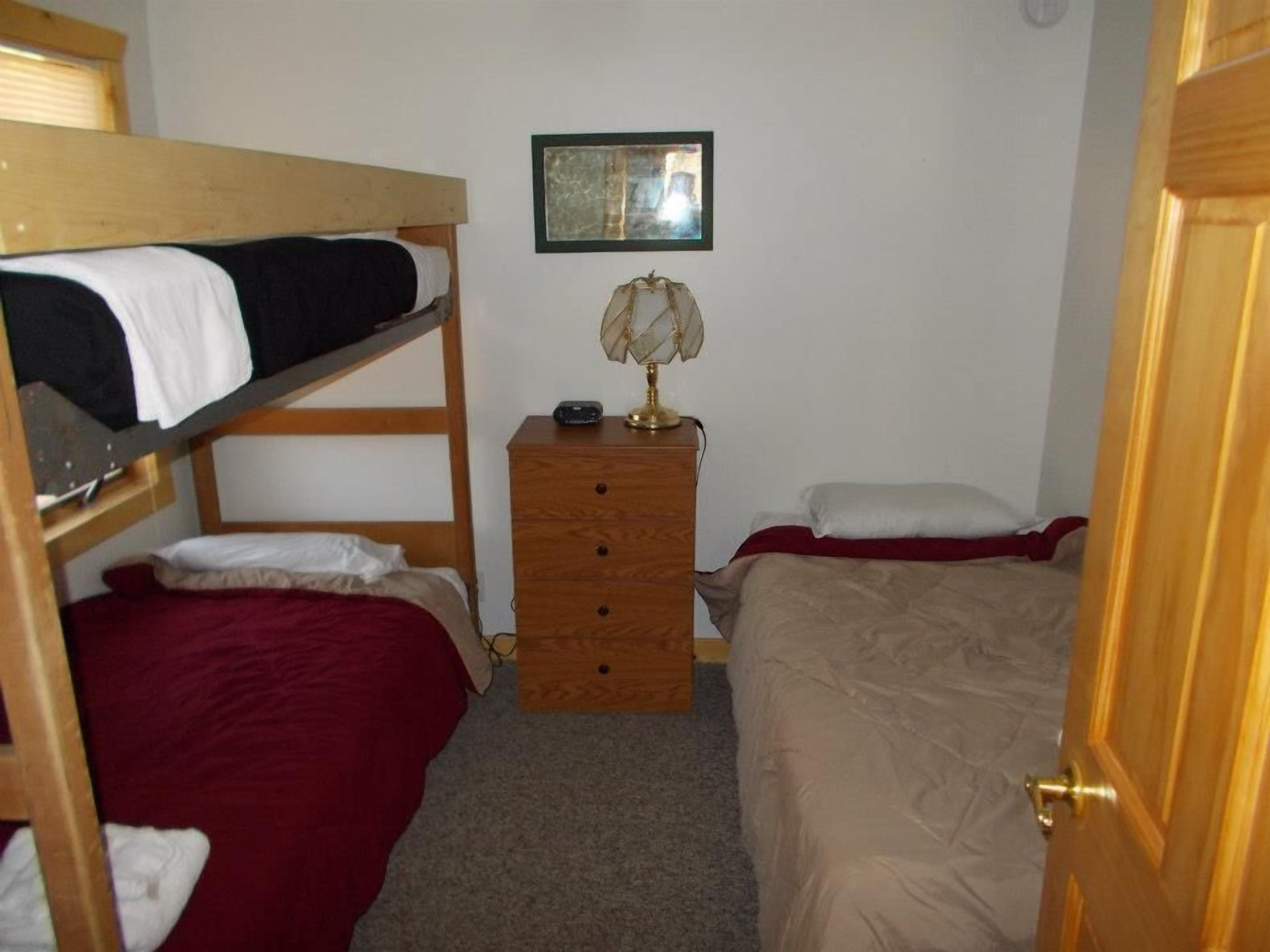A dormitory room with bunk beds, a wooden dresser, a lamp, and a framed picture on the wall.
