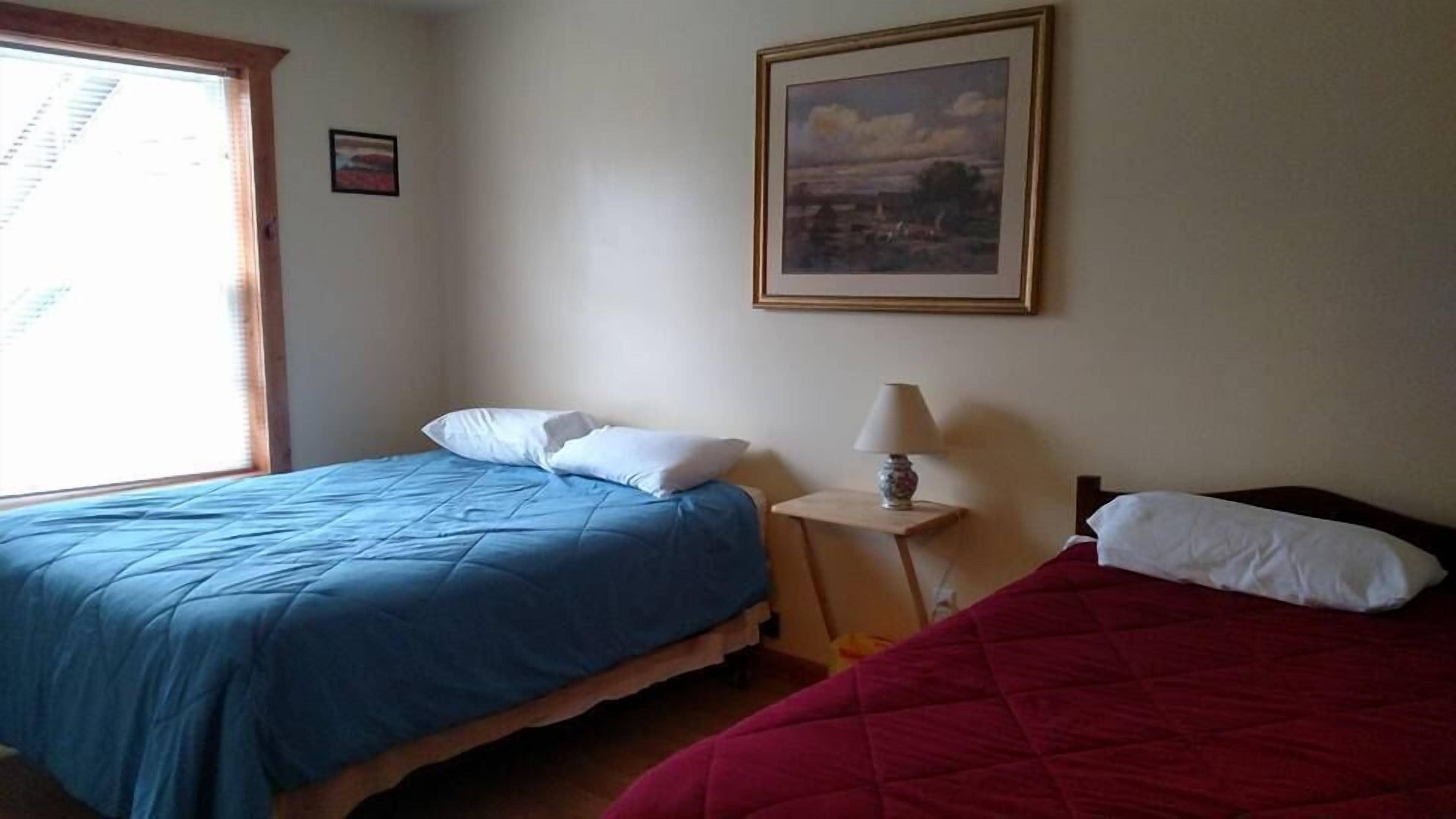 A room with two beds, one with a blue bedspread and the other with a red bedspread, a small table with a lamp, and a framed picture on the wall.