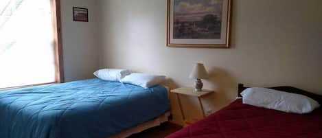 Ein Zimmer mit zwei Betten, eines mit blauer Bettdecke und das andere mit roter Bettdecke, einem kleinen Tisch mit einer Lampe und einem gerahmten Bild an der Wand.