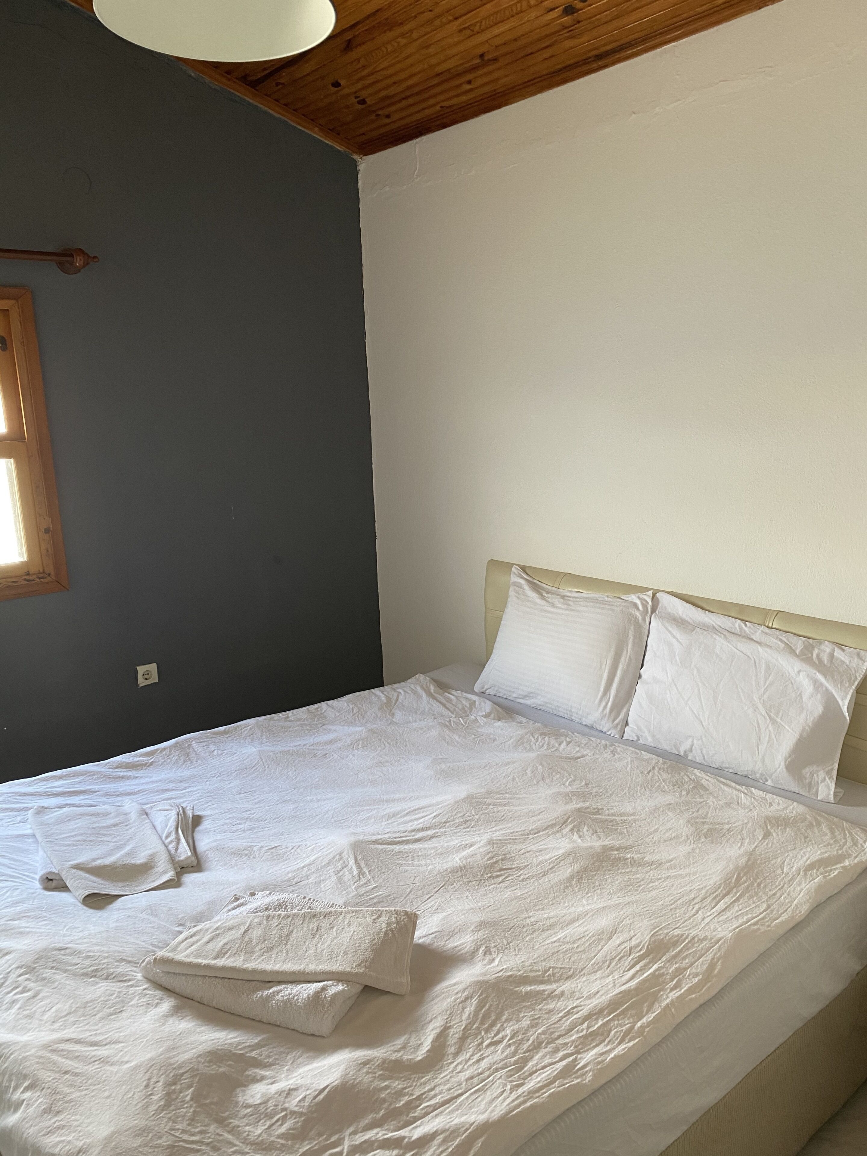 Una cama bien tendida con sábanas y almohadas blancas, un cabecero de madera y una pared gris con una ventana.