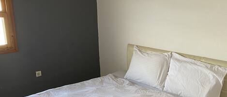 Una cama bien tendida con sábanas y almohadas blancas, un cabecero de madera y una pared gris con una ventana.