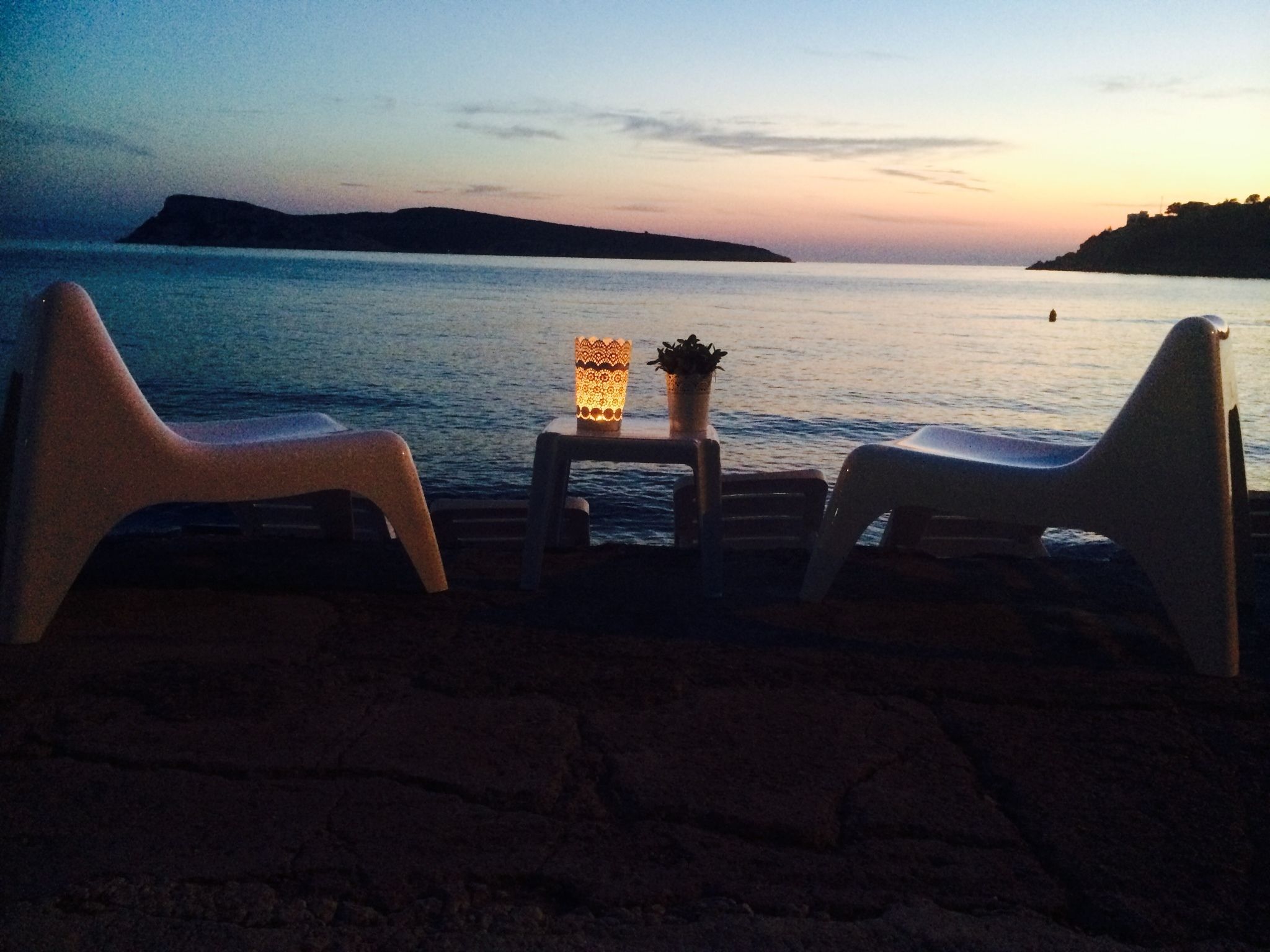 Un cadre idyllique, au bord de la plage, avec deux chaises longues blanches donnant sur une mer calme et une île lointaine. Une petite table, ornée d’une lampe décorative et d’une plante en pot, se trouve entre les chaises.