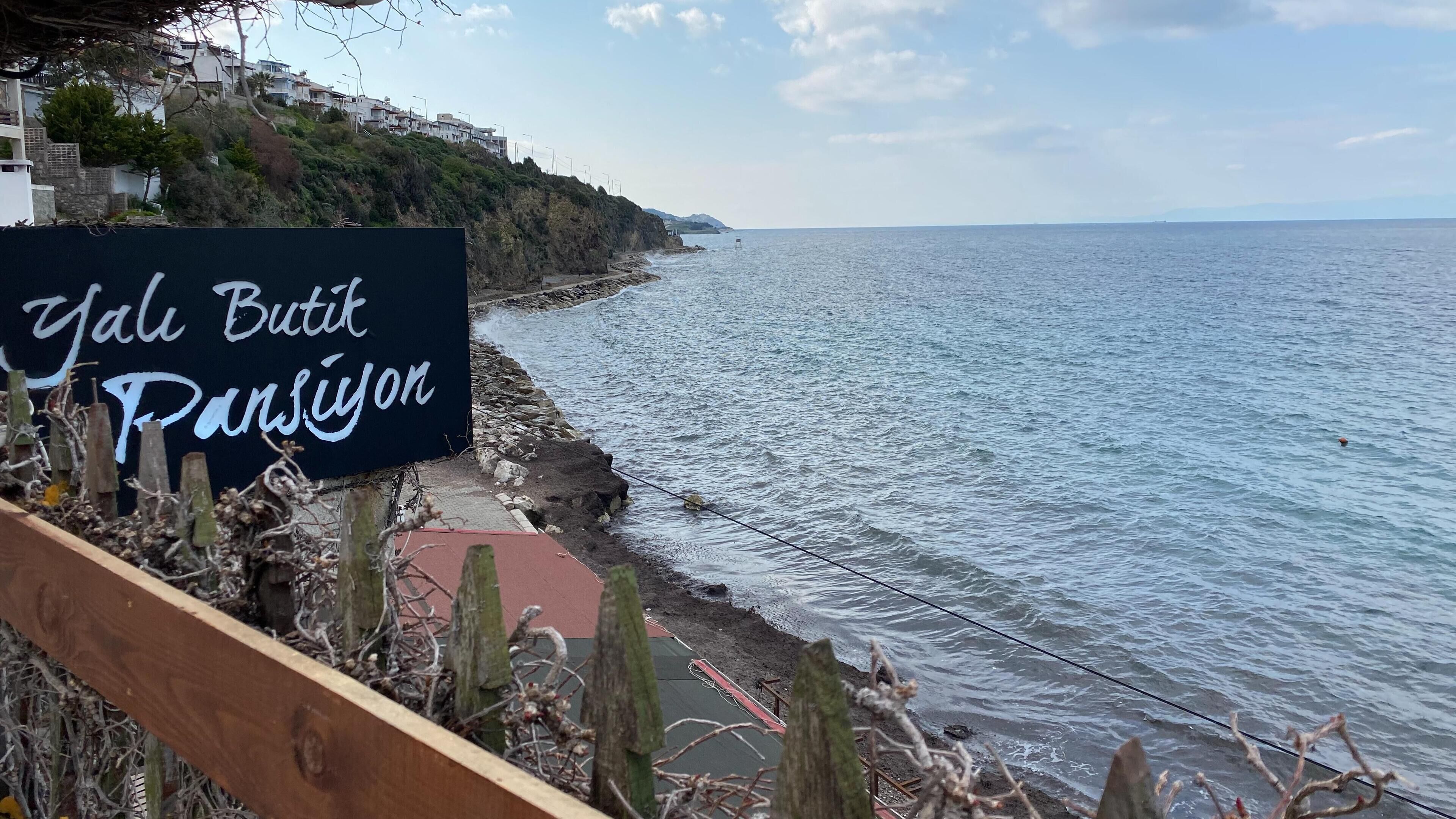A coastal view with a sign reading "Yal Butik Pansiyon" indicating a shop or establishment.