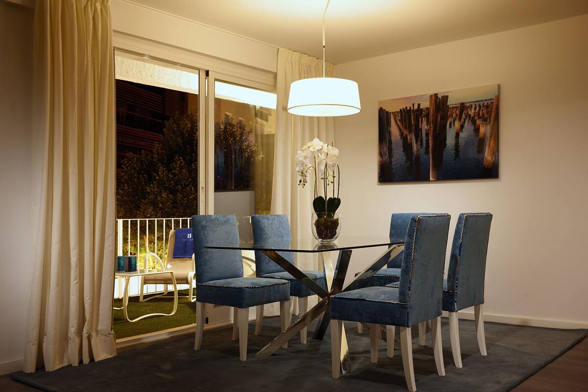 Un comedor con una mesa de vidrio, sillas tapizadas en azul, un florero con flores blancas y un cuadro en la pared.