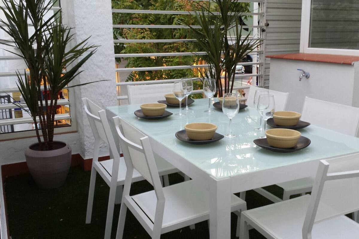 Mesa con cubierta de vidrio dispuesta para comer, con sillas blancas, rodeada de plantas en macetas.