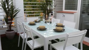 Mesa con cubierta de vidrio dispuesta para comer, con sillas blancas, rodeada de plantas en macetas.