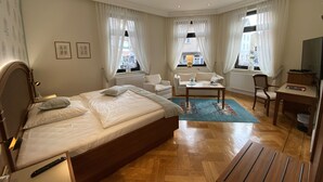 Habitación de hotel con una cama grande, un sofá, una mesita, una silla, un televisor y una ventana con cortinas.