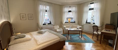 A hotel room with a large bed, a sofa, a small table, a chair, a TV, and a window with curtains.