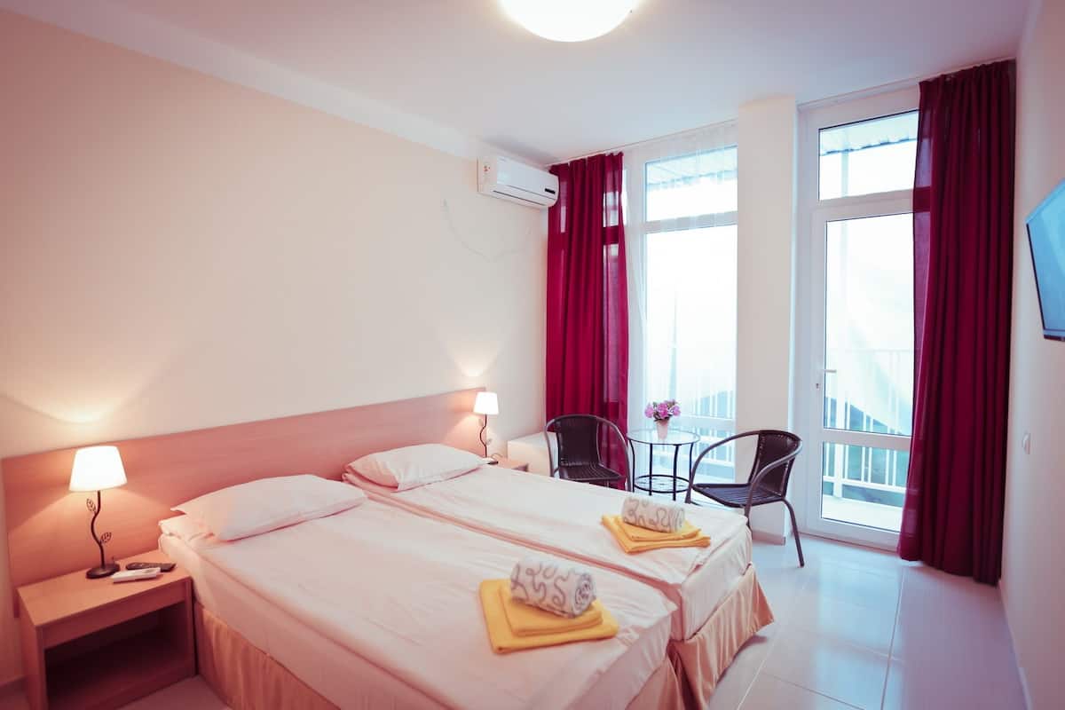 A hotel room with two beds, a desk, a chair, a TV, and a balcony with red curtains.