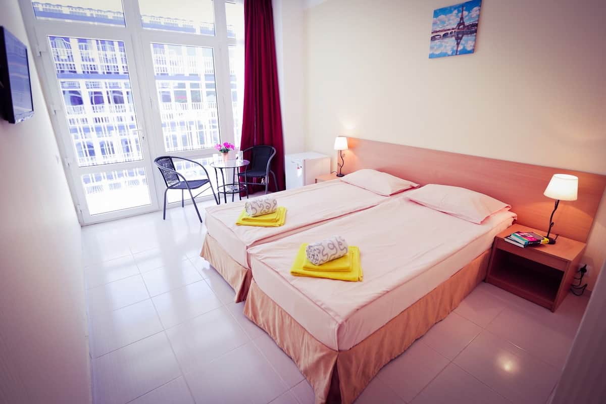 A hotel room with two beds, a desk, a chair, a window with a view of buildings, and a painting on the wall.