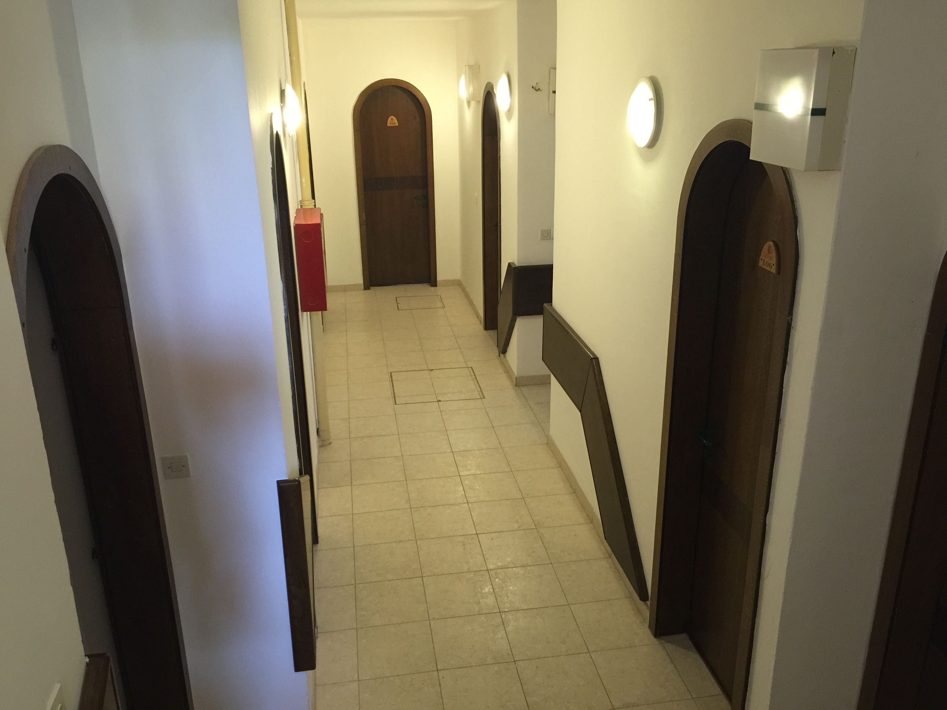 Un pasillo con suelo de baldosas, puertas de madera y luminarias fijadas a la pared.