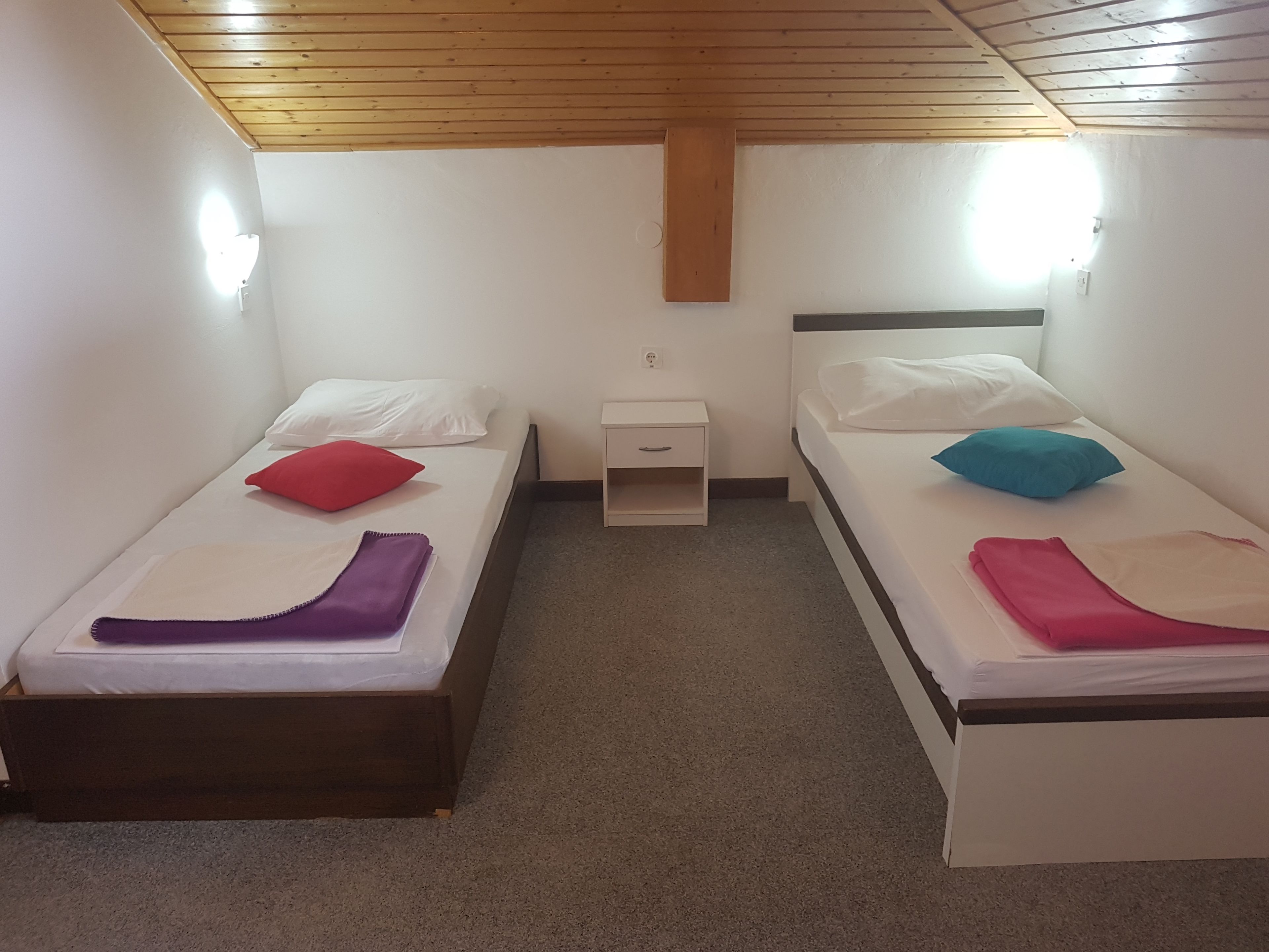 A room with two single beds, each with a pillow and a blanket. There is a small bedside table between the beds. The room has white walls and a wooden ceiling.