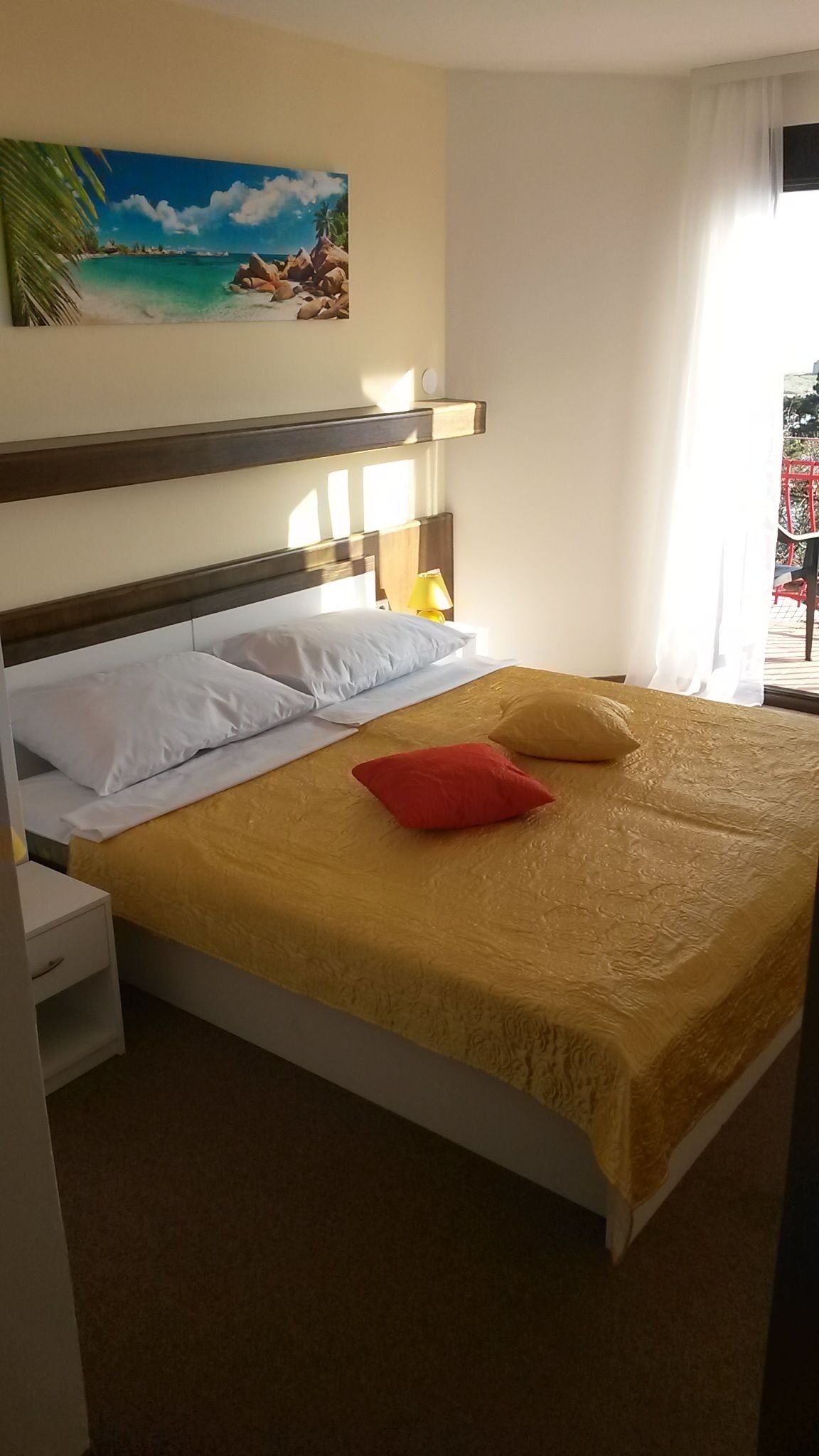 Ein ordentlich gemachtes Bett mit weißen und roten Kissen, einer gelben Tagesdecke und einem Bild einer Strandlandschaft an der Wand.
