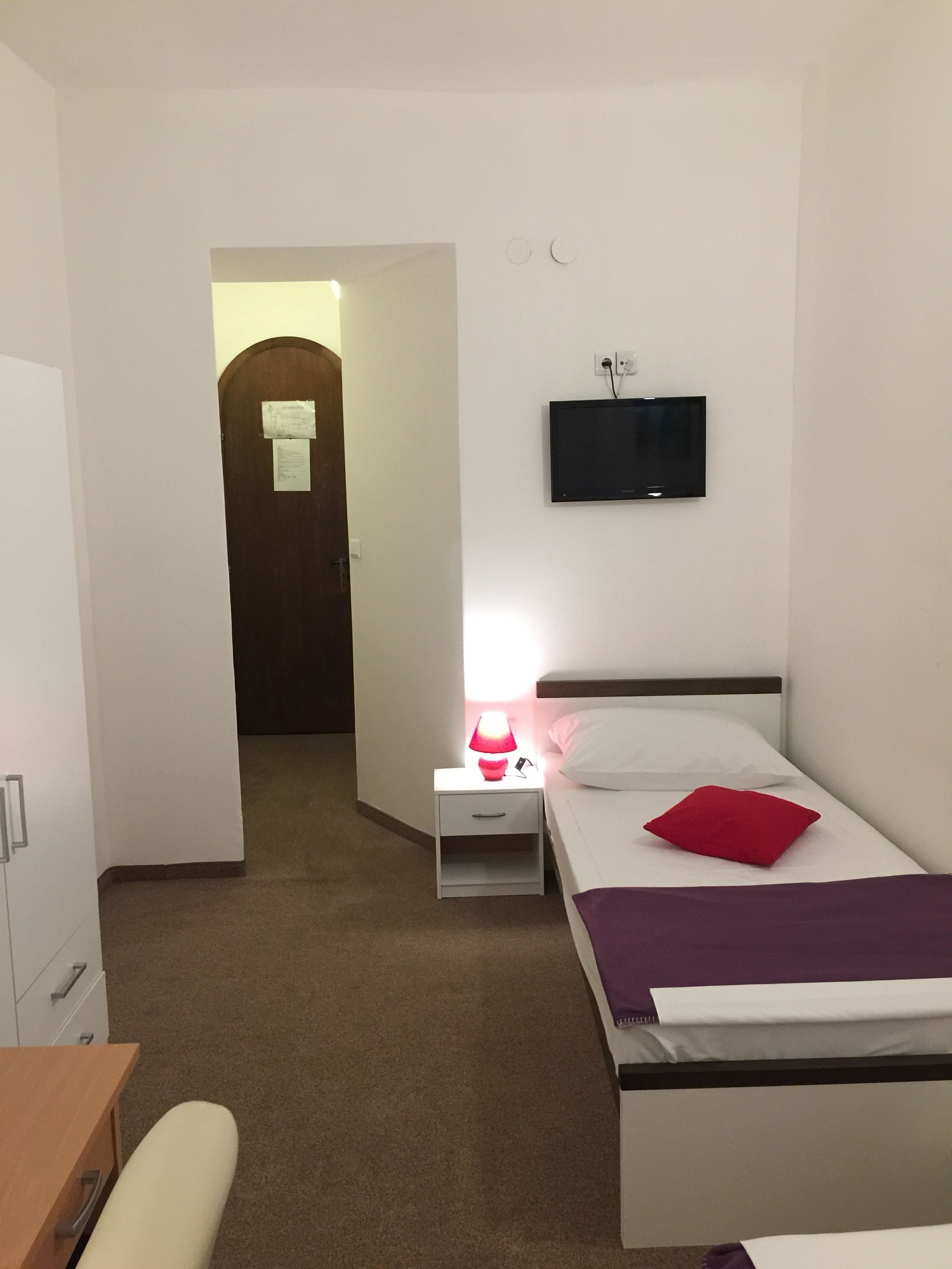 A hotel room with a bed, a bedside table with a lamp, a TV mounted on the wall, and a door leading to another room.