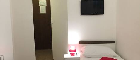 A hotel room with a bed, a bedside table with a lamp, a TV mounted on the wall, and a door leading to another room.