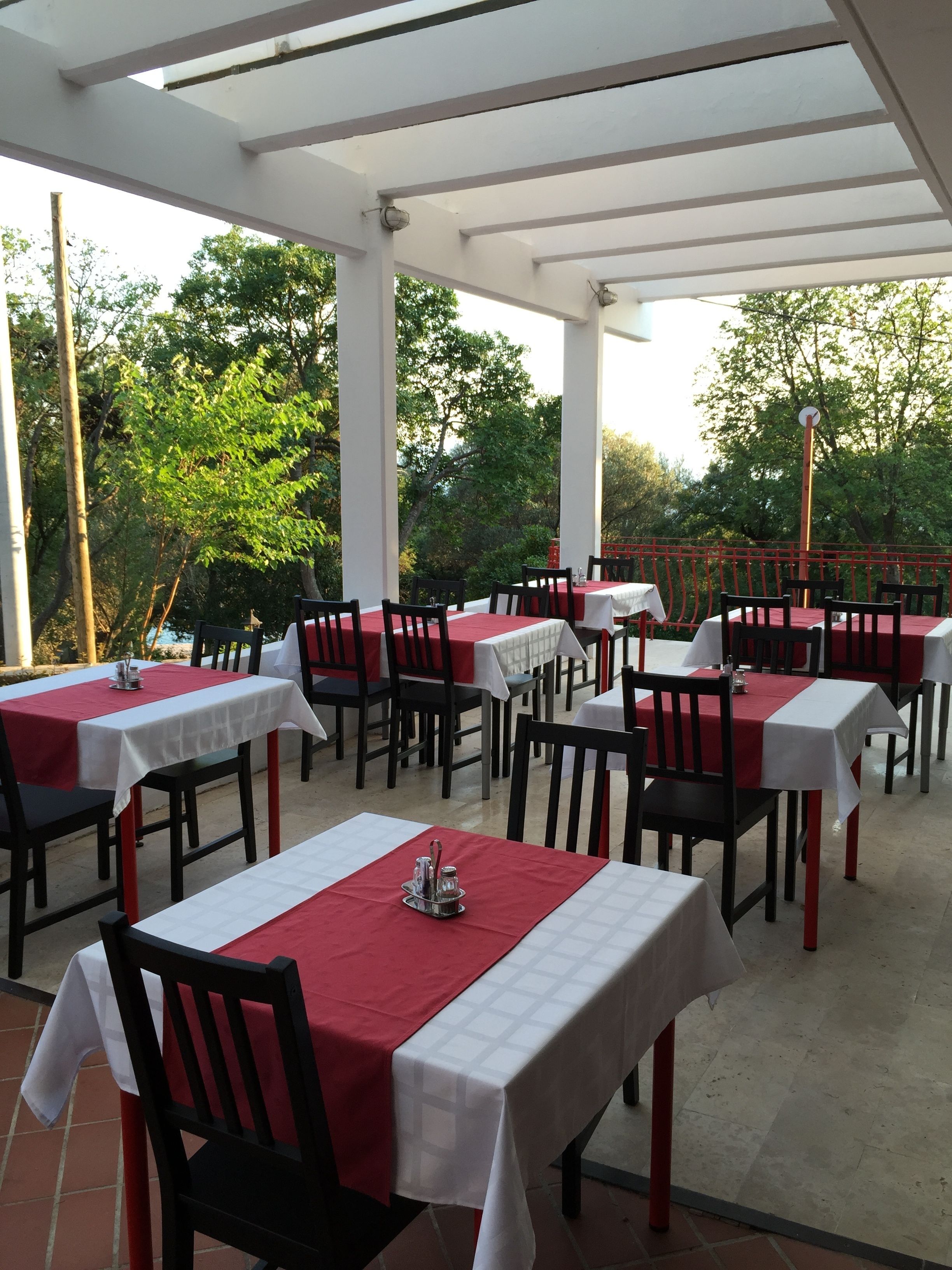 An outdoor dining area with tables and chairs, covered by a white pergola, surrounded by trees.