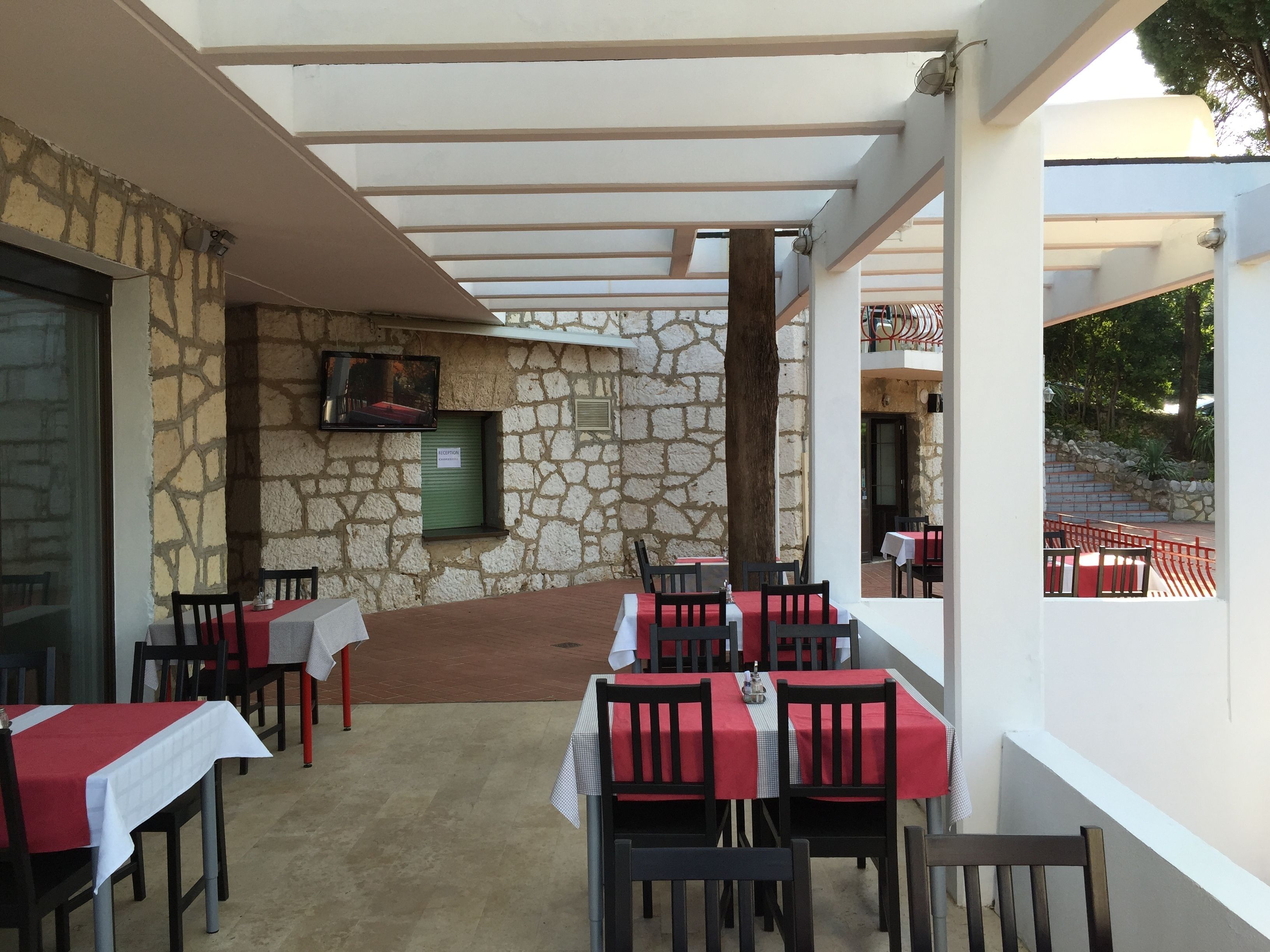 An outdoor dining area with stone walls, tables with red and white tablecloths, and a TV mounted on the wall.