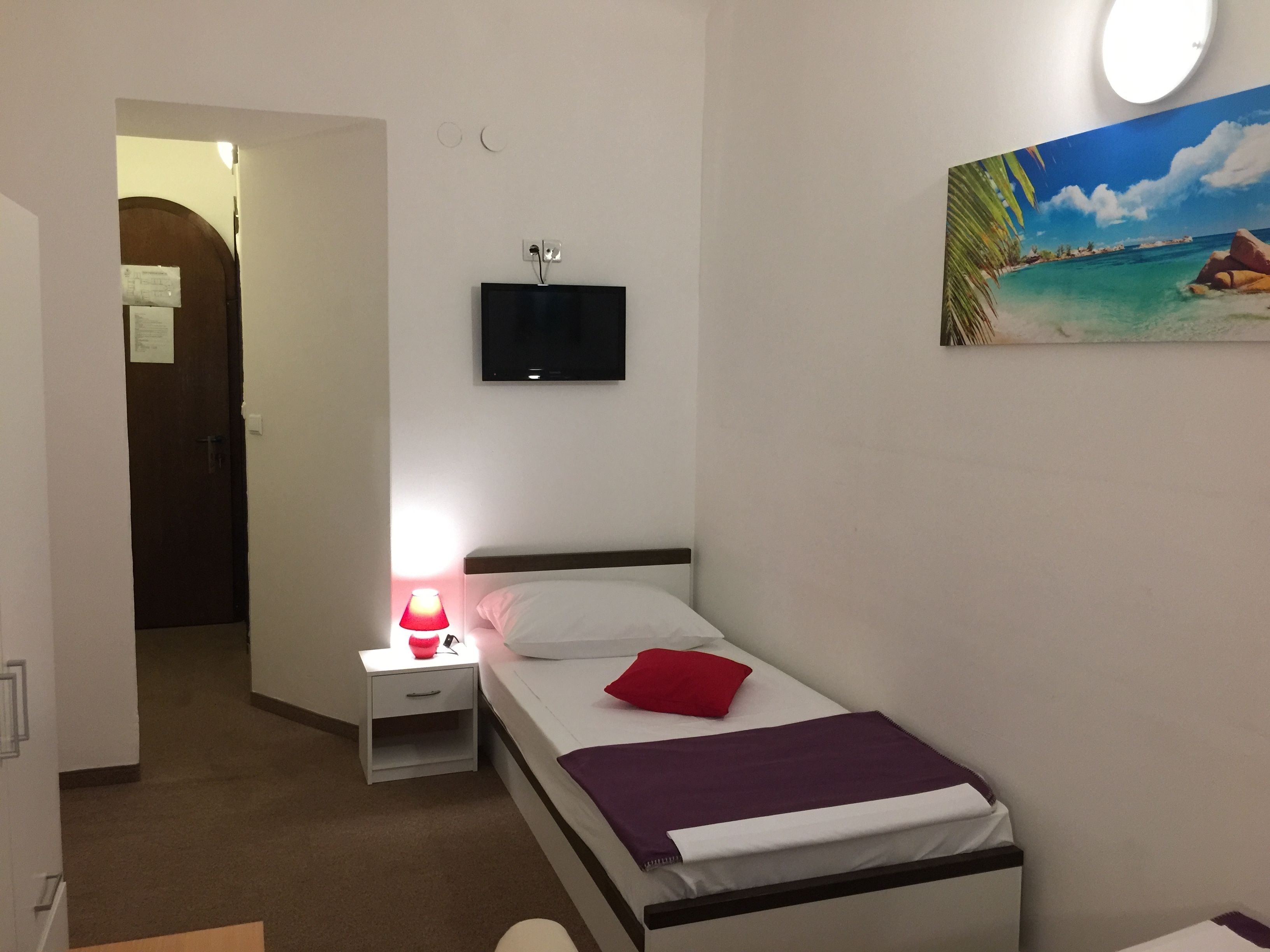 A small, neatly arranged bedroom with a single bed, a nightstand, a TV mounted on the wall, and a painting of a beach scene.