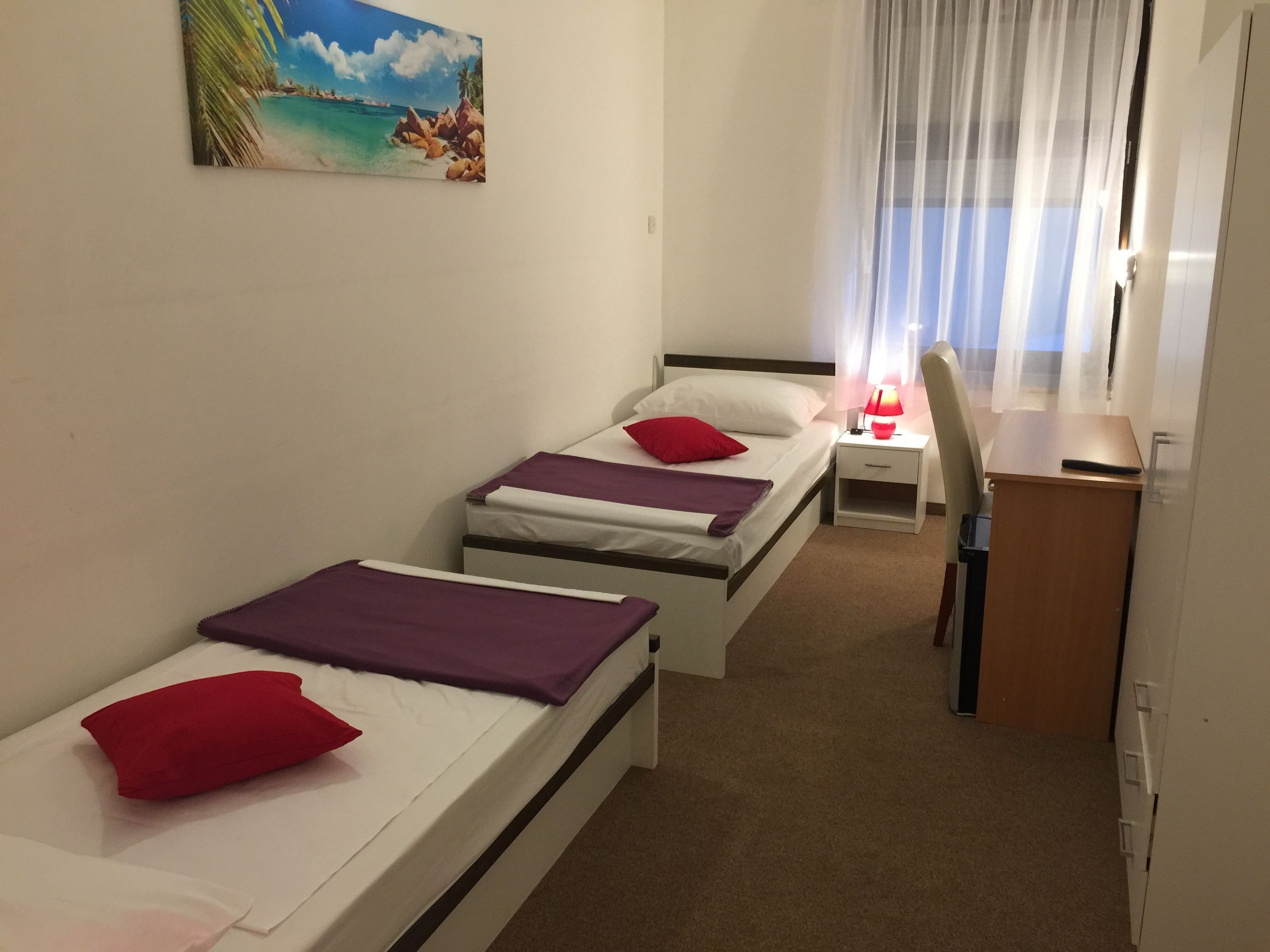 Hotellihuone, jossa on kaksi erillistä sänkyä, työpöytä, tuoli ja maalaus, jossa on kuvattuna rantamaisema.