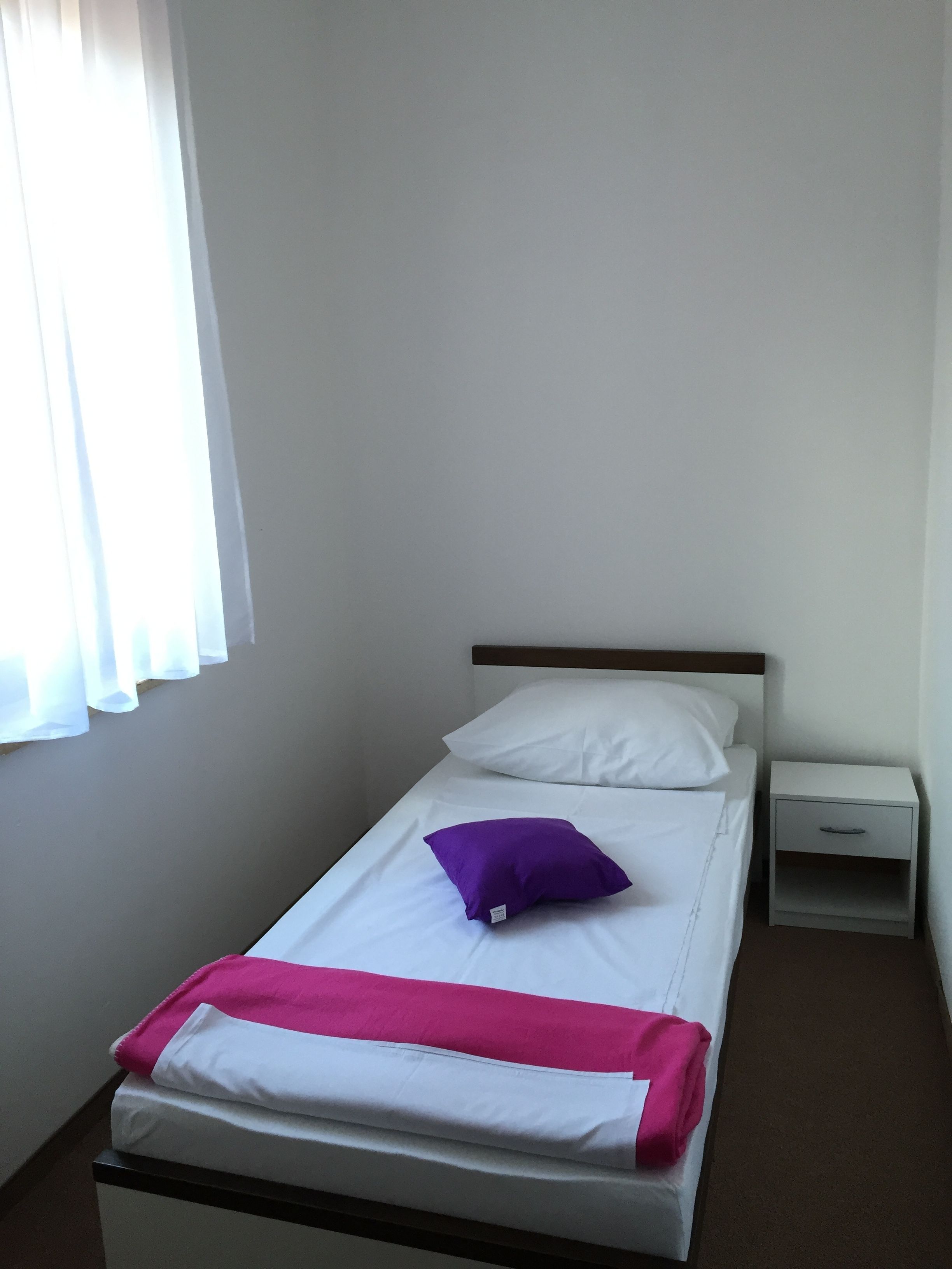 A single bed with a white mattress, a purple pillow, and a pink blanket. There is a white nightstand with a drawer to the right of the bed.