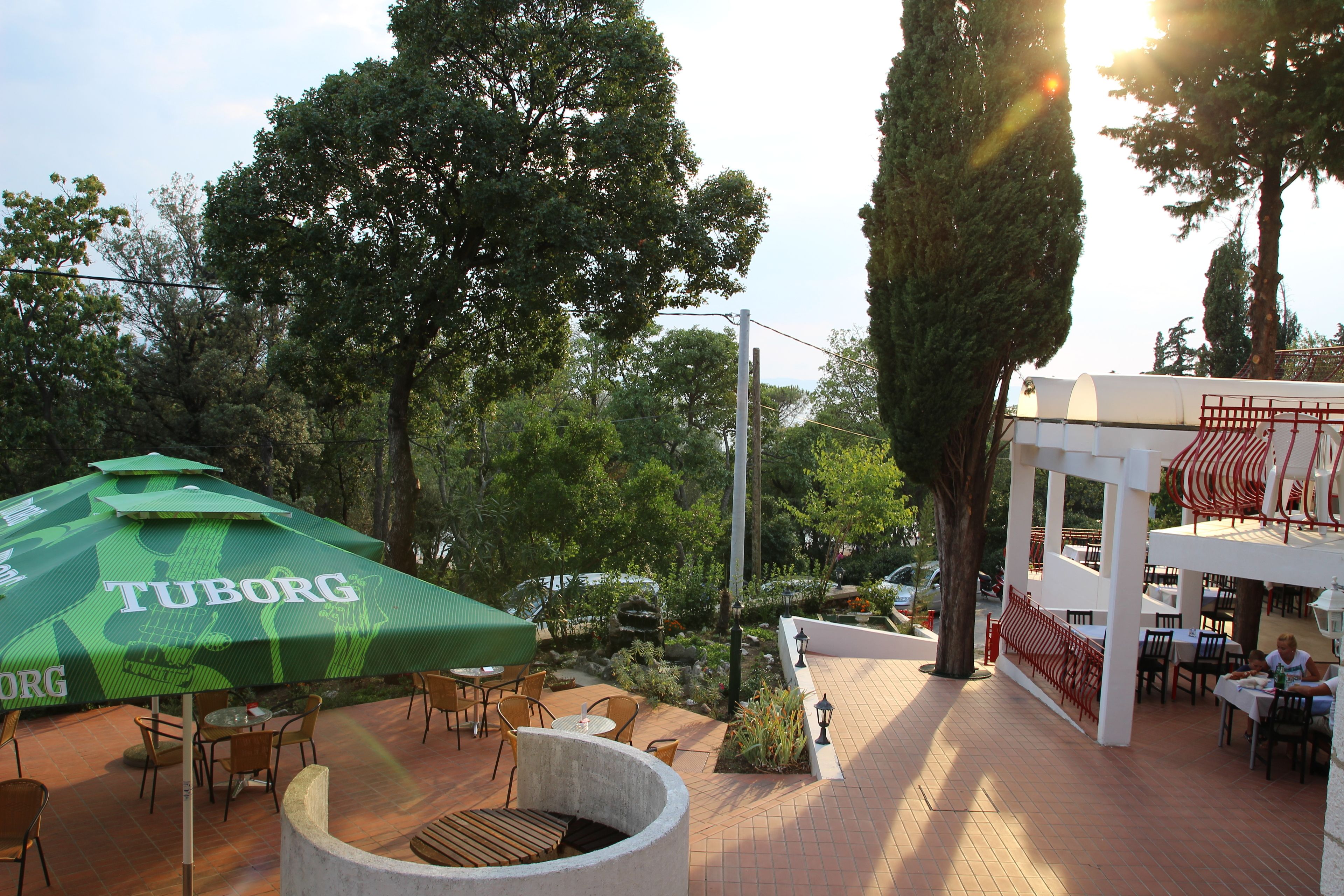 Un patio al aire libre con mesas y sillas, un paraguas verde y un edificio blanco con detalles rojos.