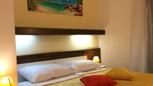 A neatly made bed with a yellow bedspread, white pillows, and a red accent pillow. There is a painting of a beach scene above the bed, and a nightstand with a lamp to the left. - Hotel Ruža (Novi Vinodolski)