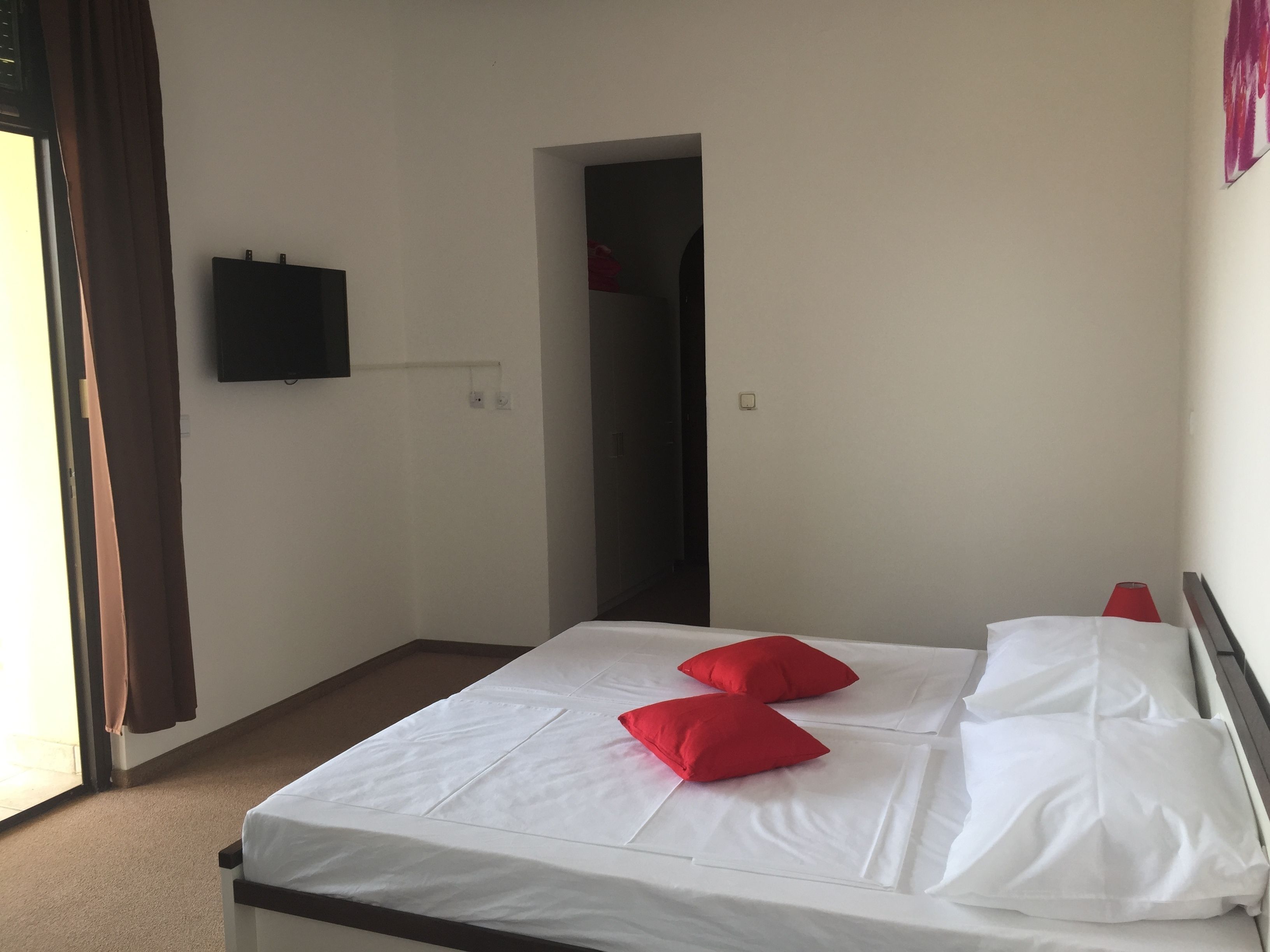 A hotel room with a bed, two red pillows, a television on the wall, and a door leading to another room.