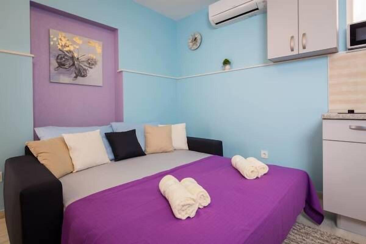 A bedroom with a bed, pillows, a wall clock, an air conditioner, and a small shelf with a plant.