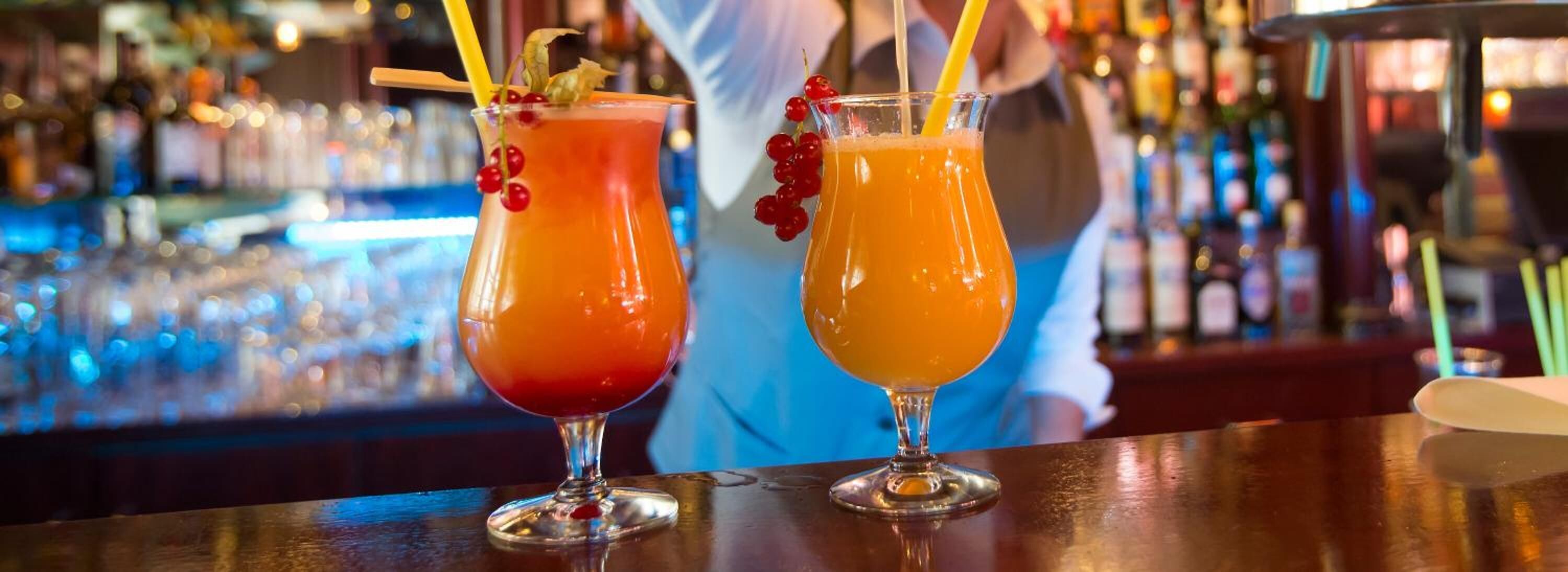Two cocktails on a bar counter with a blurred background of bottles.