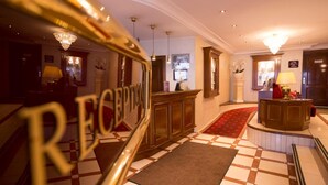 Un hall d’hôtel comprenant un comptoir d’enregistrement, un bureau de réception, un tapis rouge et un lustre.