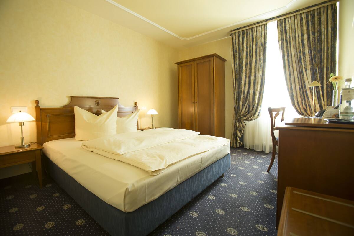 Quarto de hotel com cama, duas luminárias de cabeceira, guarda-roupa de madeira, escrivaninha com luminária e janela com cortinas.