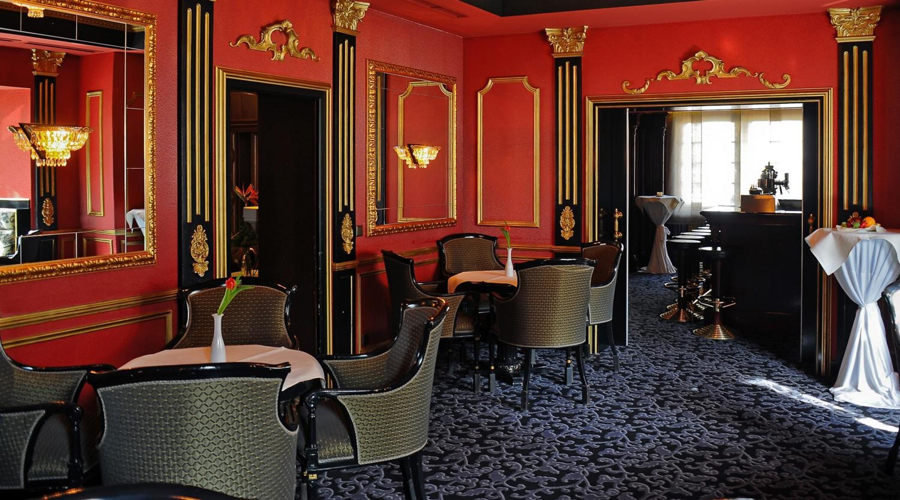 Hotelkamer met rode wanden, gouden accenten en een kamerbreed tapijt. Meerdere stoelen en een tafel met een bloevas.