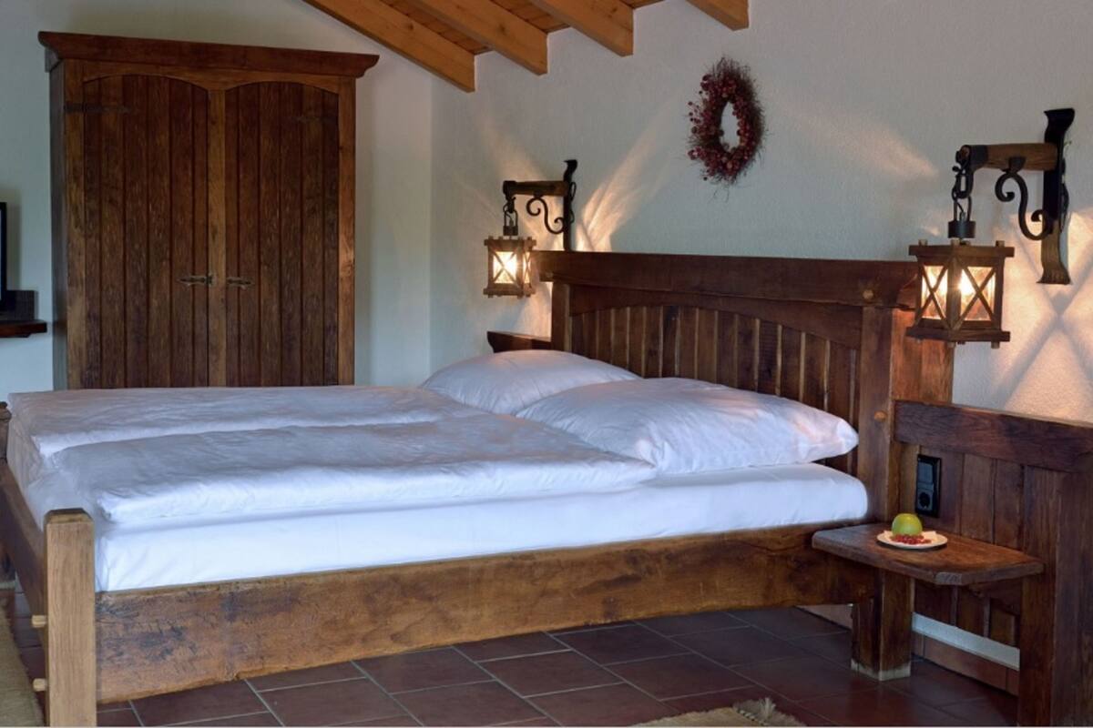 Uma cama de casal com cabeceira de madeira, duas luminárias na parede, um criado-mudo de madeira com uma travessa de fruta e um guarda-roupa de madeira.