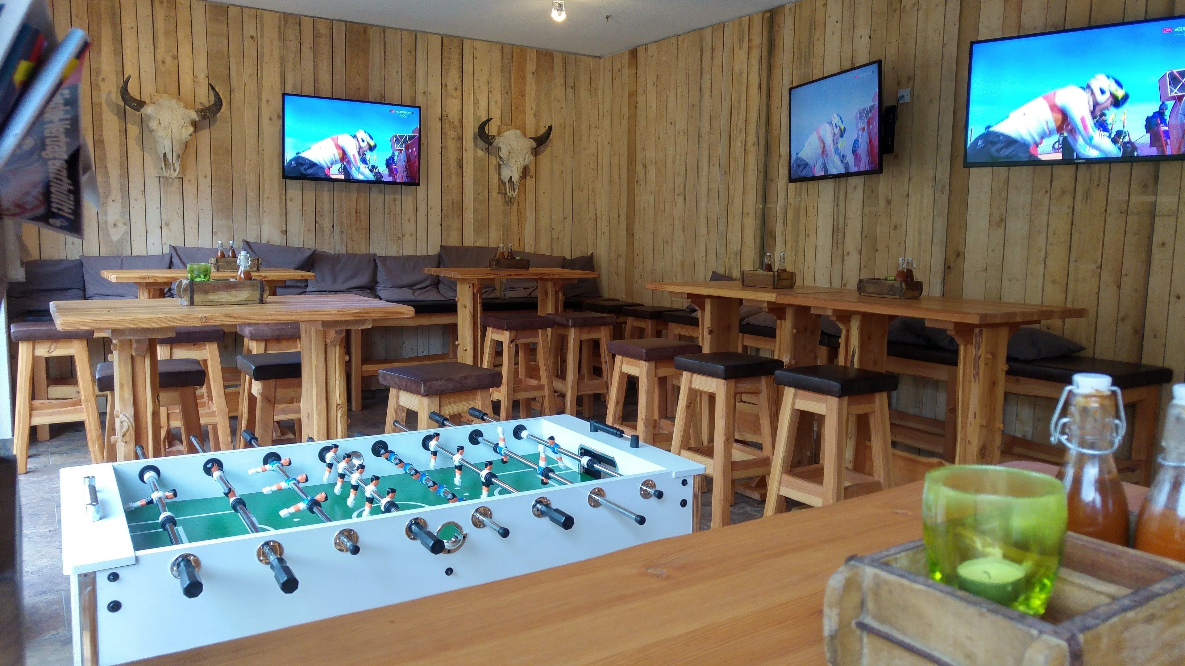 A wooden-floored bar area with a foosball table, multiple seating options, and two mounted televisions.